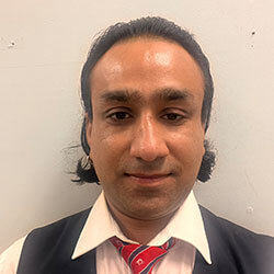 Portrait of a man in a navy blazer, white shirt, and red striped tie, facing the camera against a light wall.