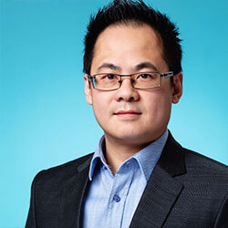 Professional headshot of a man wearing glasses, a blue shirt, and a dark blazer against a teal background.