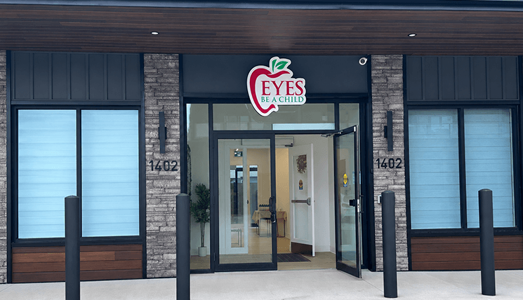 Front entrance of an 'Eyes Be a Child' clinic, glass doors open, with a brick and wood façade and the address 1402 on both sides.